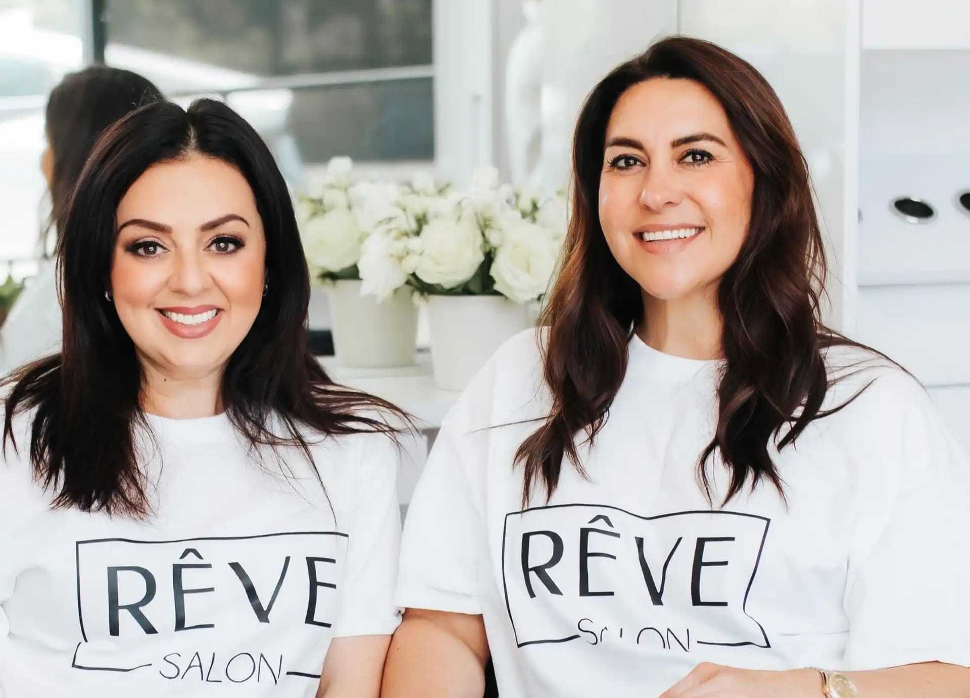 Two women smiling, wearing "Rêve Salon" shirts, with white flowers in the background.